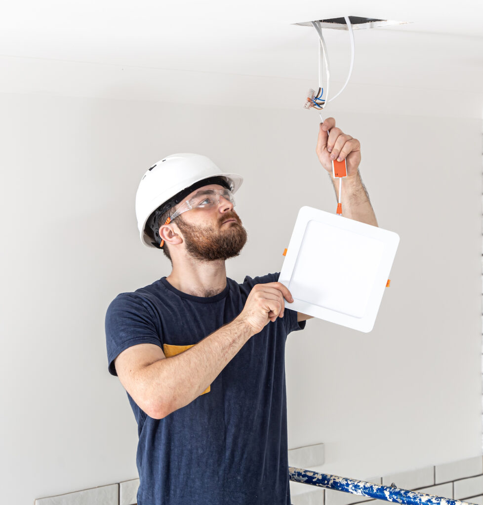 Electrician Builder with beard worker in a white helmet at work, installation of lamps at height. Professional in overalls with a drill on the background of the repair site.